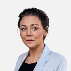 A person with dark hair smiles slightly, wearing a light blue blazer over a black top. The background is plain white.