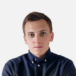 A person with short dark hair wearing a dark collared shirt is looking directly at the camera against a plain white background.