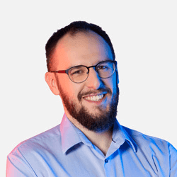 Smiling man with glasses and a beard, wearing a light blue shirt, standing in front of a plain background with blue and red lighting.