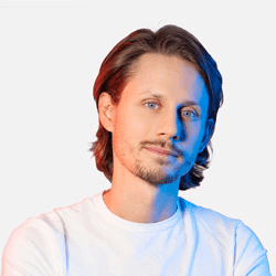A person with shoulder-length hair and a short beard is looking at the camera with a slight smile. They are wearing a white T-shirt, and there are colorful blue and red lights on their face. The background is plain white.