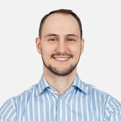 A man with short hair and a beard smiles at the camera. He is wearing a light blue and white striped shirt. The background is plain white.