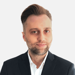 A man with short hair and a beard, wearing a dark suit and white shirt, stands against a plain background. He is looking at the camera with a slight smile.