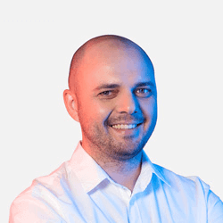 A smiling man with a bald head and a beard wearing a white shirt. The lighting is blue and red, creating a gradient effect on his face. He stands against a plain light gray background.