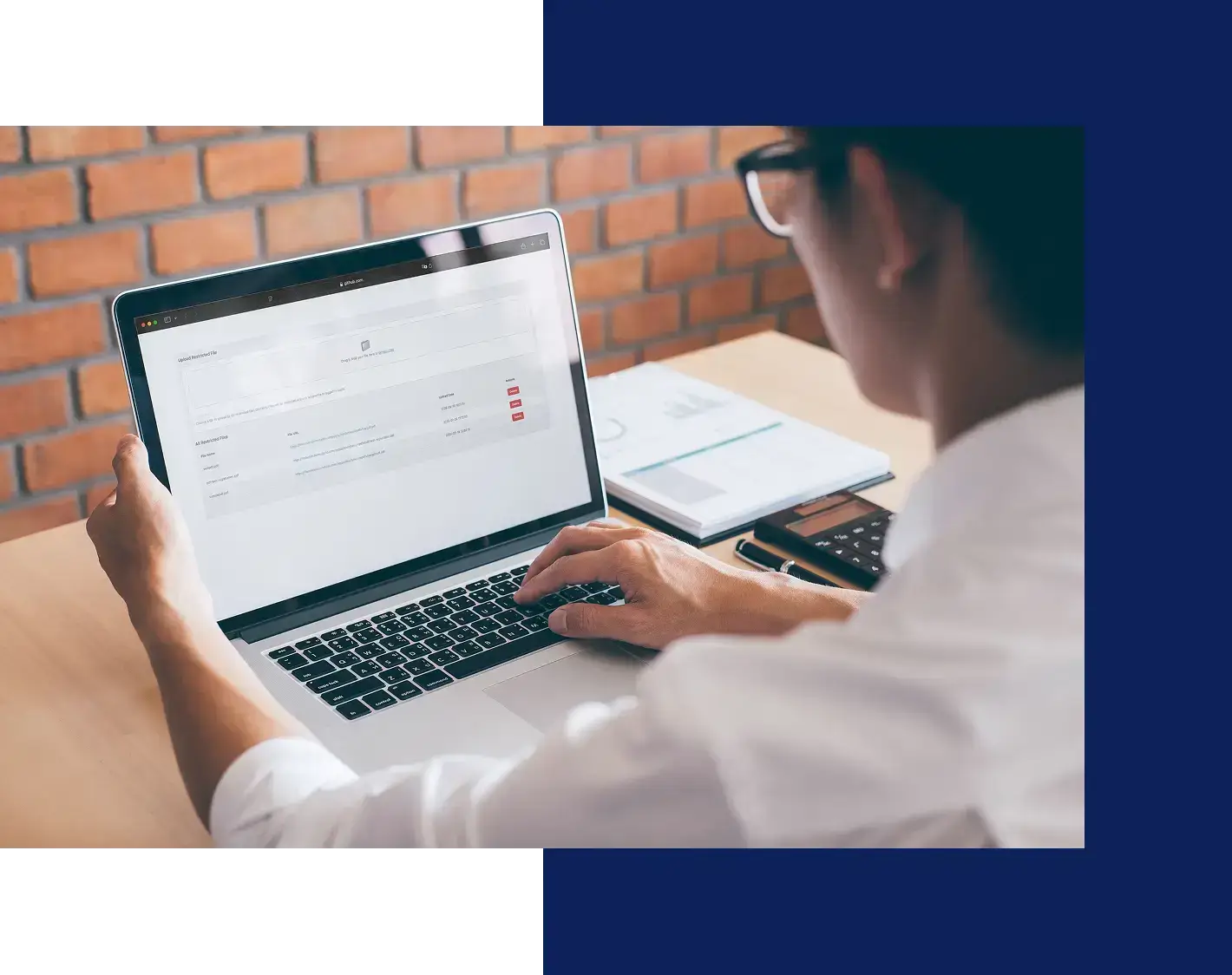 A person wearing glasses and a white shirt uses a laptop at a desk with papers and a calculator, shown from behind. The computer screen displays a web page with a form or data entry interface.